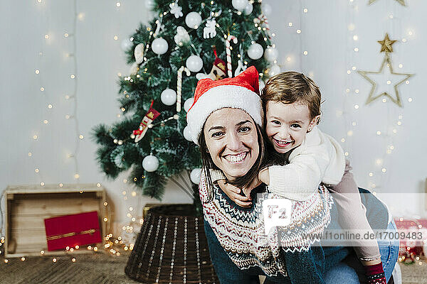 Lächelnde Mutter,  die ihre niedliche Tochter zu Hause während der Weihnachtszeit huckepack nimmt