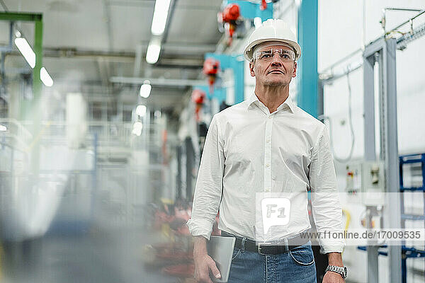 Selbstbewusster reifer Manager mit Brille und Schutzhelm  der in einer Fabrik nachdenkt