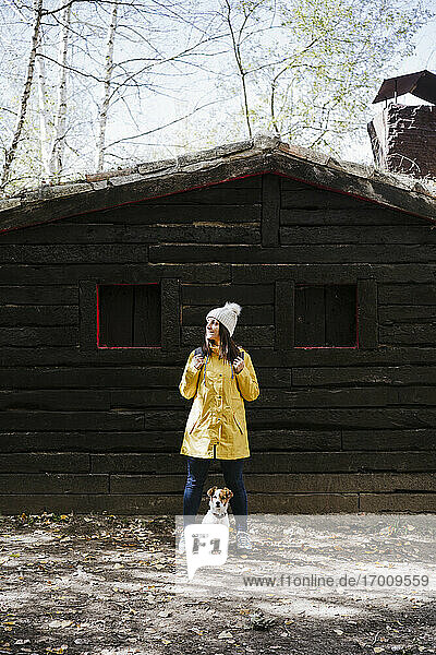 Frau in gelbem Regenmantel steht mit Hund vor einer Blockhütte