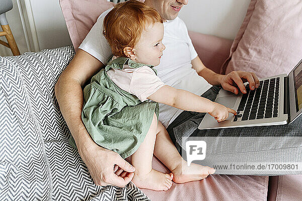 Man with baby daughter working on laptop on sofa