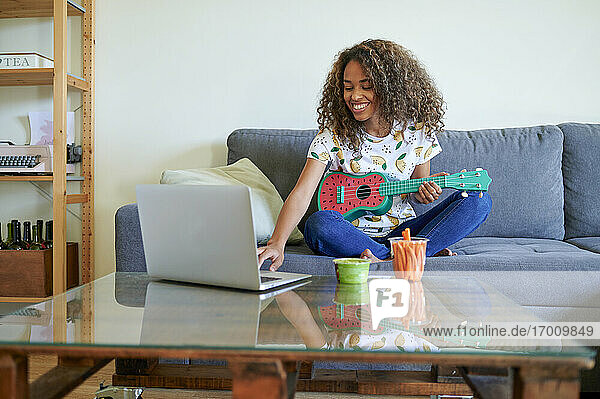 Lächelnde Frau  die am Laptop im Wohnzimmer Ukulele spielen lernt