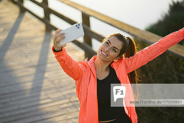 Lächelnde junge Frau  die ein Selfie mit ihrem Mobiltelefon macht  während sie auf einer Fußgängerbrücke steht