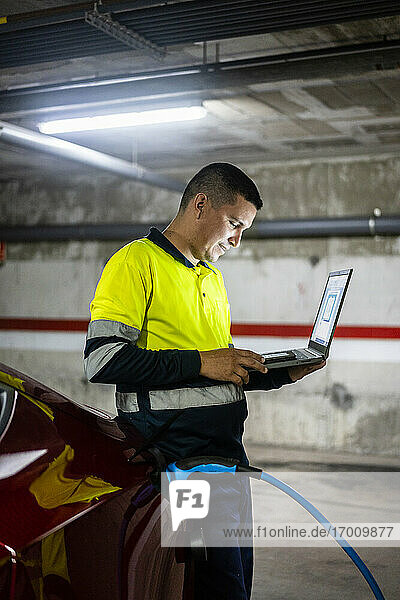 Lächelnder männlicher Techniker mit Laptop  der ein Mobiltelefon an einem Elektroauto in einer Autowerkstatt benutzt