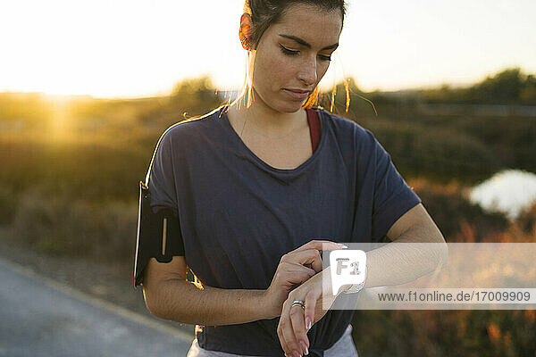 Junge Frau  die eine intelligente Uhr benutzt  während sie bei Sonnenuntergang vor der Landschaft steht