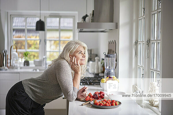 Nachdenkliche Rentnerin lehnt sich mit Erdbeeren in der Wohnung an den Küchentisch