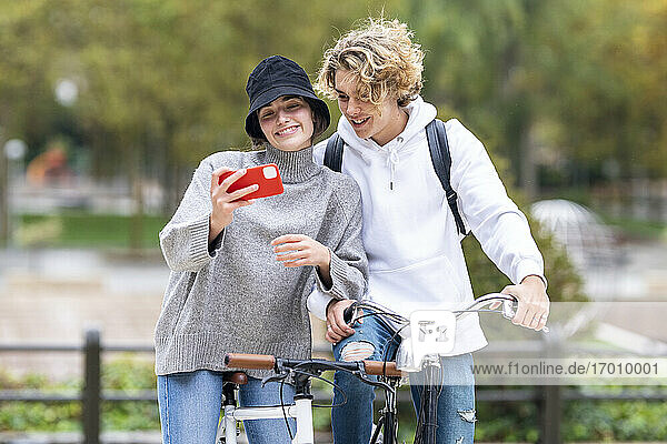 Smiling woman taking selfie standing by male friend with bicycle in park
