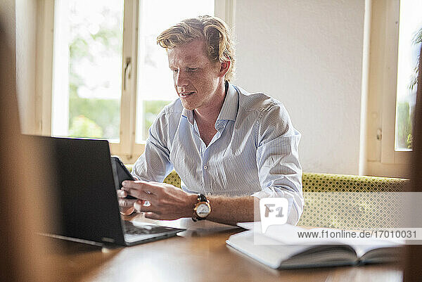 Geschäftsmann  der ein Smartphone benutzt  während er zu Hause am Laptop arbeitet