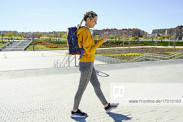 Weibliche Sportlerin mit Rucksack  die beim Gehen auf dem Fußweg ein Mobiltelefon benutzt