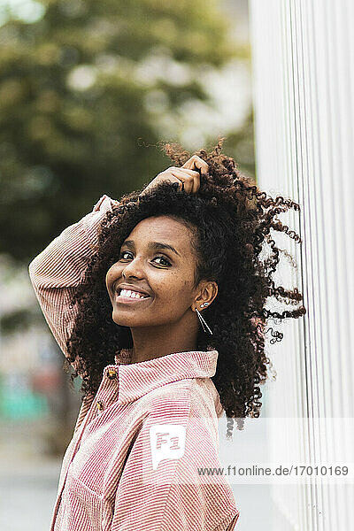 Afro woman smiling with hand in hair while standing in city