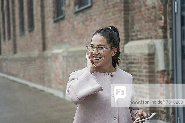 Lächelnde Geschäftsfrau  die ihr Smartphone in der Hand hält und gegen ein Gebäude blickt