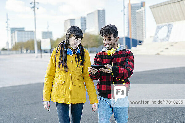 Lächelndes Paar mit Kopfhörern und Mobiltelefon beim Gehen auf der Straße