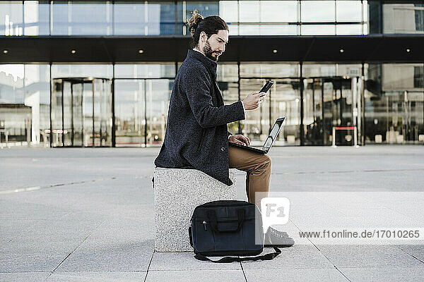 Geschäftsmann mit Laptop  der ein Mobiltelefon benutzt  während er auf einer Bank sitzt