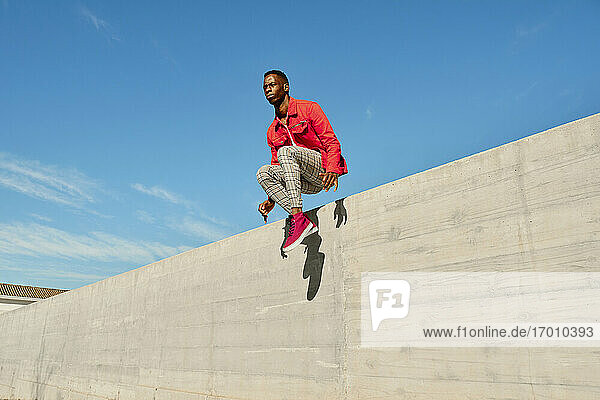 Junger Mann in roter Jacke und karierter Hose springt von einer Betonmauer