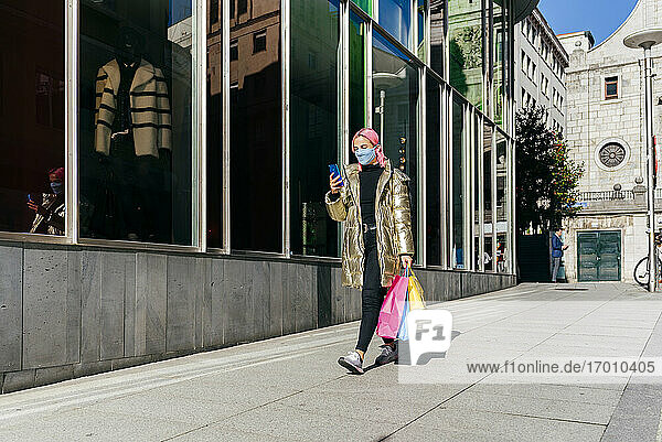 Frau mit Gesichtsschutz  die ein Mobiltelefon benutzt  während sie mit Einkaufstaschen auf dem Gehweg geht