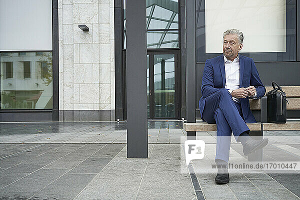 Älterer Geschäftsmann  der mit gekreuzten Beinen an einer Bushaltestelle in der Stadt sitzt und wartet