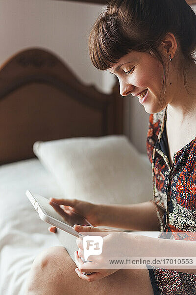 Lächelnde Frau  die auf dem Bett sitzend ein Video auf einem digitalen Tablet betrachtet