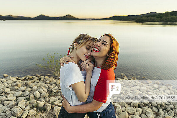 Glückliche Frau  die ihre Freundin umarmt  während sie bei Sonnenuntergang am See steht