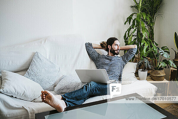 Mann mit Händen hinter dem Kopf sitzt mit Laptop auf dem Sofa zu Hause