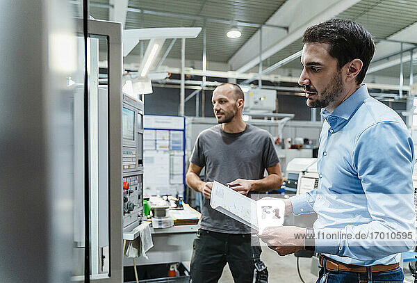 Männlicher Ingenieur mit Dokument  der Maschinen untersucht  während er mit einem Wartungsarbeiter im Hintergrund in einer Fabrik steht