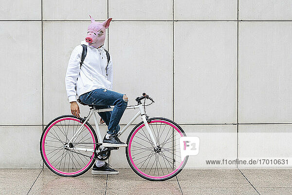 Young man sitting on bicycle with pig mask against wall on footpath