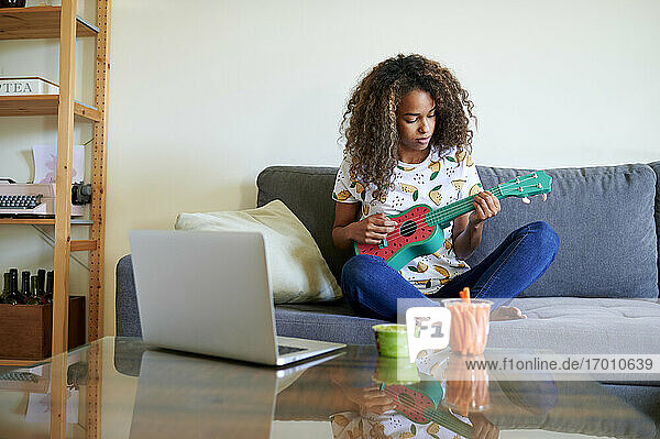 Afro junge Frau mit Laptop spielt Ukulele im Wohnzimmer
