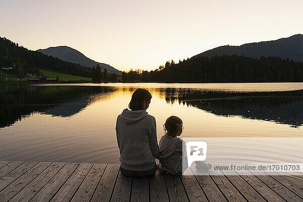 Mutter und kleine Tochter sitzen in der Abenddämmerung zusammen am Ende eines Stegs am Seeufer