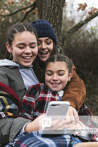 Drei lächelnde Schwestern schauen auf ihr Smartphone in einer Herbstlandschaft