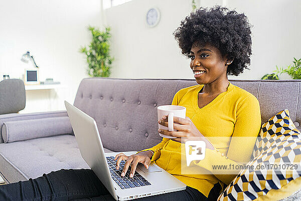 Lächelnde Frau mit Kaffeetasse und Laptop  während sie zu Hause auf dem Sofa sitzt