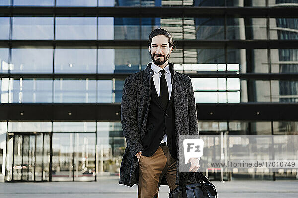 Männlicher Unternehmer mit Händen in der Tasche  der eine Aktentasche trägt  während er vor einem modernen Gebäude steht