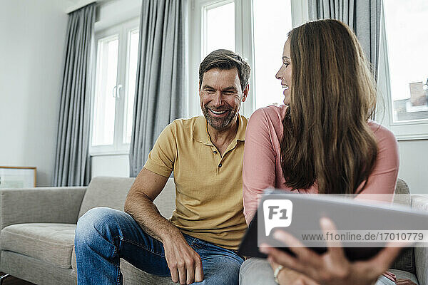 Reife Frau mit digitalem Tablet  die einen glücklichen Mann ansieht  während sie auf dem Sofa in einer Wohnung sitzt
