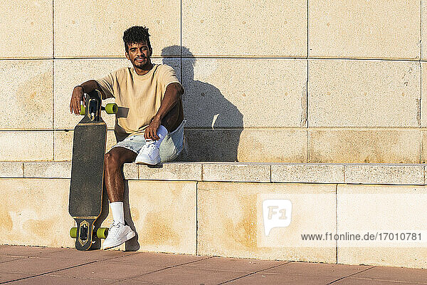 Lächelnder junger Mann mit Skateboard  der bei Sonnenuntergang auf einer Stützmauer sitzt