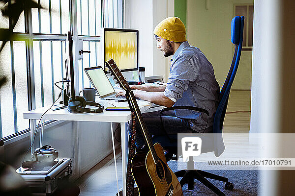 Musiker arbeitet am Laptop  während er im Studio an der Gitarre sitzt