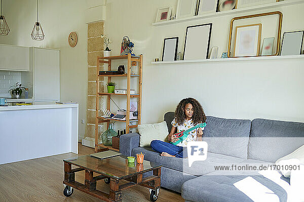 Junge Afro-Frau spielt Ukulele  während sie zu Hause auf dem Sofa sitzt