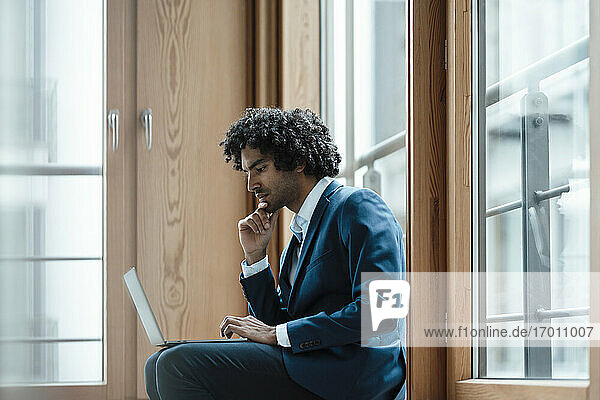 Junger männlicher Unternehmer  der einen Laptop benutzt  während er am Arbeitsplatz am Fenster sitzt