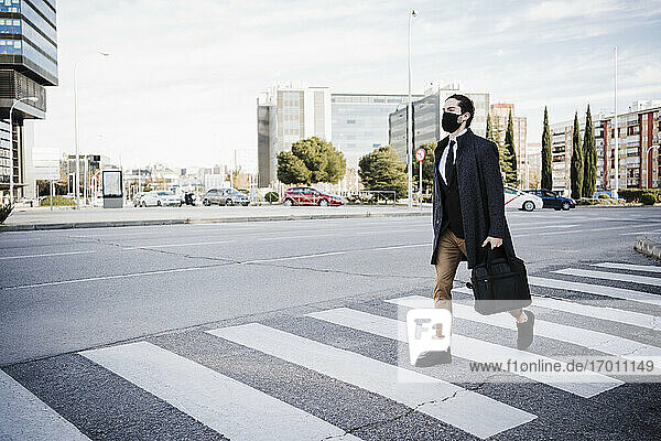 Geschäftsmann mit Jacke und Gesichtsmaske  der eine Aktentasche hält  während er auf einer Straße in der Stadt geht