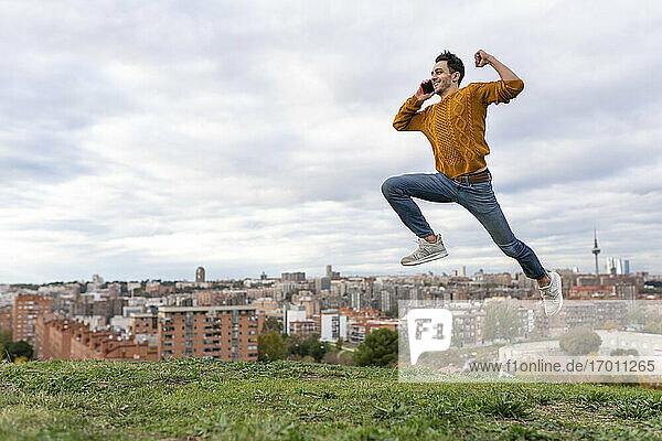 Aufgeregter junger Mann  der mit seinem Handy spricht  während er über einen Hügel vor einer Stadtlandschaft springt