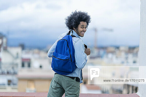 Fröhlicher junger Mann mit blauem Rucksack auf einer Terrasse in der Stadt