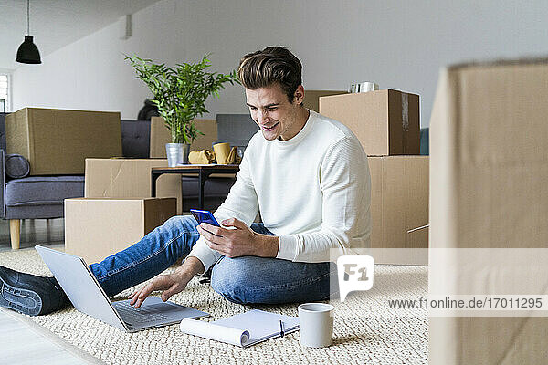 Lächelnder Mann mit Laptop  der ein Smartphone benutzt  während er im Wohnzimmer seines neuen Hauses sitzt