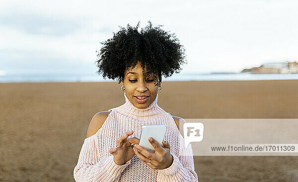 Frau mit Afro-Haar  die ein Mobiltelefon benutzt  während sie am Strand gegen den Himmel steht