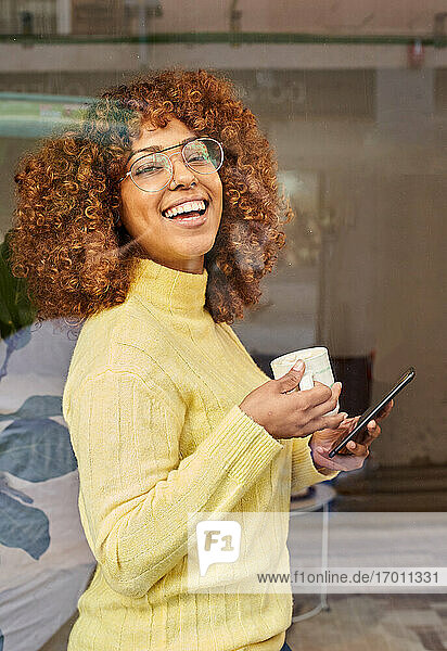 Lachende Frau mit Mobiltelefon beim Kaffee trinken hinter Glas in einem Cafe