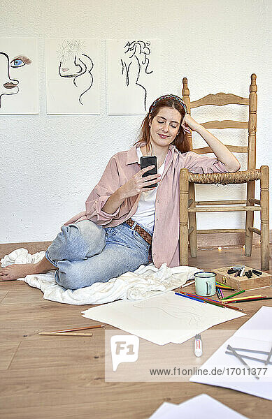 Lächelnde Künstlerin  die ein Mobiltelefon benutzt  während sie sich zu Hause an einen Stuhl lehnt