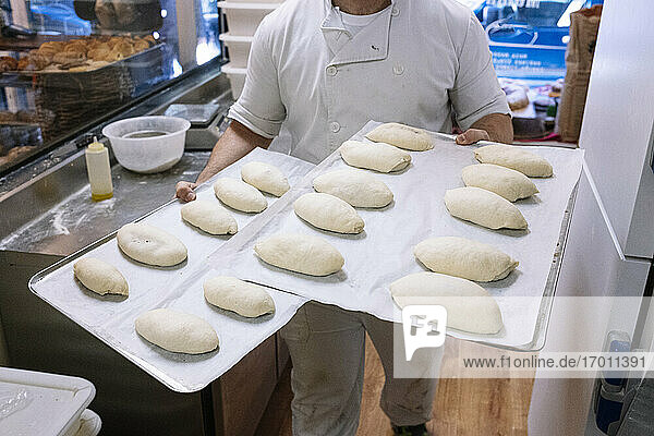 Männlicher Bäcker geht mit einem Backblech  das mit Mehlteig gefüllt ist  in der Küche einer Bäckerei