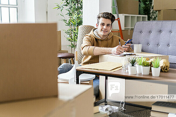 Lächelnder Mann mit Kugelschreiber  der ein Mobiltelefon über einer Box in einer neuen Wohnung hält