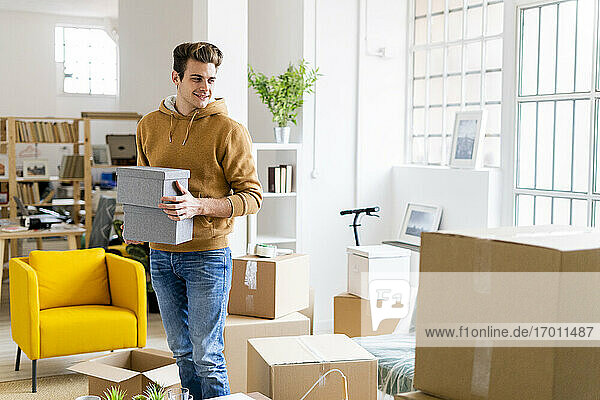 Lächelnder junger Mann mit Kisten in der Hand in seiner neuen Loftwohnung
