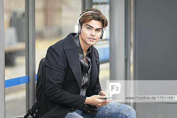 Hübscher junger Mann hört Musik  während er an der Bushaltestelle sitzt