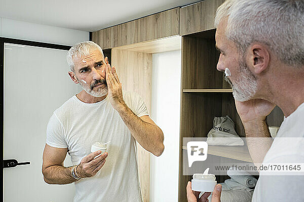 Mature man applying face cream while looking in mirror at bathroom