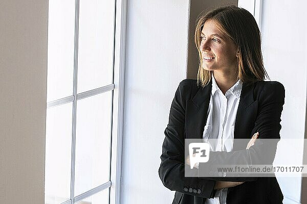 Nachdenkliche junge Geschäftsfrau mit verschränkten Armen beim Blick durch das Fenster zu Hause