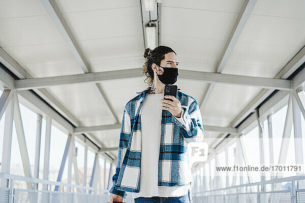 Junger Mann mit Gesichtsmaske  der wegschaut  während er auf einer Brücke steht und ein Mobiltelefon benutzt
