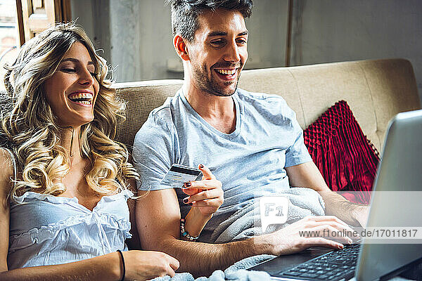 Happy young couple sitting on couch at home with credit card and laptop