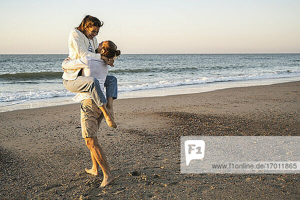 Carefree young man lifting girlfriend at beach during sunset